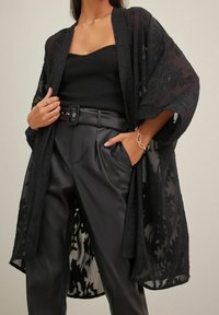 Czarny kwiecisty kimono z koronką o przezroczystej strukturze, z otwartym przodem, szerokimi rękawami, nałożone na czarny prążkowany top na ramiączkach i skórzane spodnie z wysokim stanem.