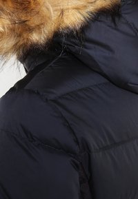 Zwarte pufferjas met een nepbontkraag. Het materiaal is glanzend en gewatteerd, met een doorgestikt ontwerp en een contrasterende textuur bij de kraag.