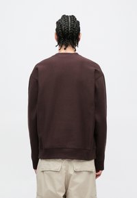 Felpa marrone con scollo rotondo e polsini a costine, caratterizzata da una vestibilità rilassata e tessuto strutturato, vista da dietro senza loghi visibili.