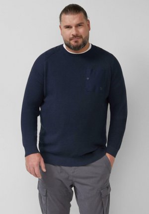 Navy niebieski sweter z prążkowanego materiału z okrągłym dekoltem, z kieszenią na piersi i długimi rękawami; noszony z szarymi spodniami cargo.
