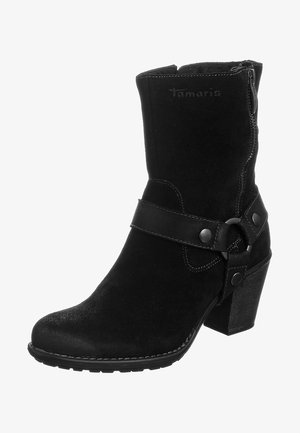 Sort ankelstøvle i ruskind med chunky hæl, sidelås, dekorativ rem med metalring og knapper samt syningsdetaljer på sålen.