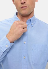 Camicia azzurro chiaro con colletto e bottoni bianchi, caratterizzata da una tasca sul petto sinistro e realizzata in tessuto strutturato.