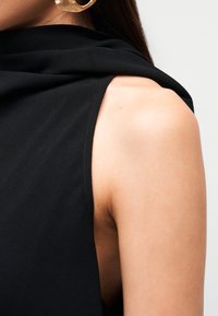 Μαύρο αμάνικο τοπ με υφασμάτινη λεπτομέρεια που drapeάρει γύρω από τον ώμο, παρουσιάζοντας ομαλή υφή και καθαρές γραμμές. Ορατό χρυσό σκουλαρίκι σε σχήμα κρίκου.