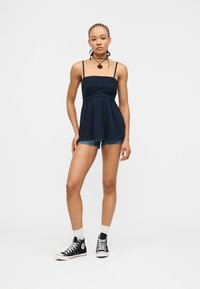 Νεαρή γυναίκα με πλεξούδες φοράει μπλε καμιζόλ, τζιν σορτς, λευκές κάλτσες και μαύρα ψηλά αθλητικά παπούτσια, στέκεται μπροστά σε απλό φόντο.