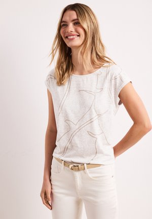 Smilende kvinde med blondt hår iført hvid bluse med abstrakt mønster og korte ærmer, beige bælte og hvide bukser, stående mod en ensfarvet baggrund.