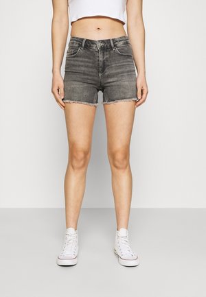 Vrouw draagt grijze denim shorts met hoge taille en rafelige zoom, een witte cropped top en witte hoge sneakers, staand tegen een effen achtergrond.
