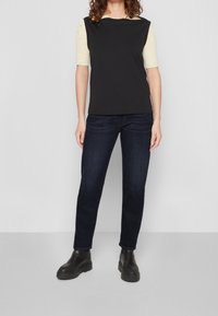 Černý top bez rukávů s texturovanou světle žlutou spodní vrstvou, doplněný tmavě modrými džíny s rovnými nohavicemi a černými robustními kotníkovými botami.