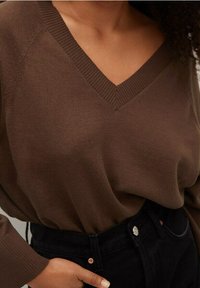 Brązowy sweter z dzianiny z dekoltem w serek i teksturowanymi detalami, zestawiony z czarnymi spodniami z wysokim stanem ozdobionymi metalowymi guzikami.