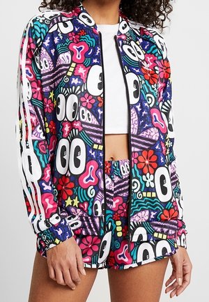 Frau trägt eine lebhaft mehrfarbige Jacke und Shorts mit auffälligen Cartoon-Augen, Blumen und abstrakten Formen auf dunklem Hintergrund.