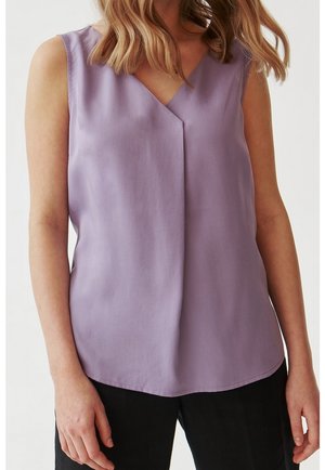 Kobieta w bezrękawniku w kolorze lawendowym, z dekoltem w kształcie litery V, i czarnych spodniach, stojąca ze swobodnie opuszczonymi ramionami.