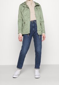 Persona che indossa un giubbotto verde chiaro, un maglione a collo alto beige, jeans blu e sneaker alte bianche, in piedi su un pavimento grigio chiaro.