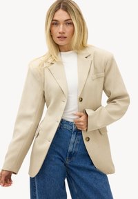 Femme blonde portant un blazer beige sur un haut blanc et un jean bleu taille haute, posant avec une main près des boutons de la veste.