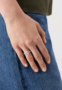 Anello d'argento con una pietra centrale ovale, circondata da pietre più piccole, montato su una fascia testurizzata. Mano resa su uno sfondo di denim.