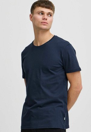 Junger Mann in einem schlichten dunkelblauen kurzärmligen T-Shirt, der nach links blickt vor einem hellgrauen Hintergrund.