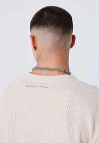 Giovane con taglio di capelli sfumati, che indossa un maglione a coste color crema e una collana in argento, rivolto di spalle su uno sfondo semplice.