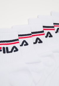 Calcetines de algodón blancos con textura de canalé, que presentan un acento a rayas en negro y rojo en la parte superior y un logo en negro.