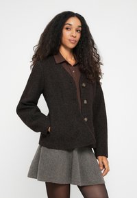 Vrouw met lang krullend haar in een donkerbruin knopen cardigan, een bruine blouse, een grijze geplooide rok en zwarte panty, tegen een witte achtergrond.