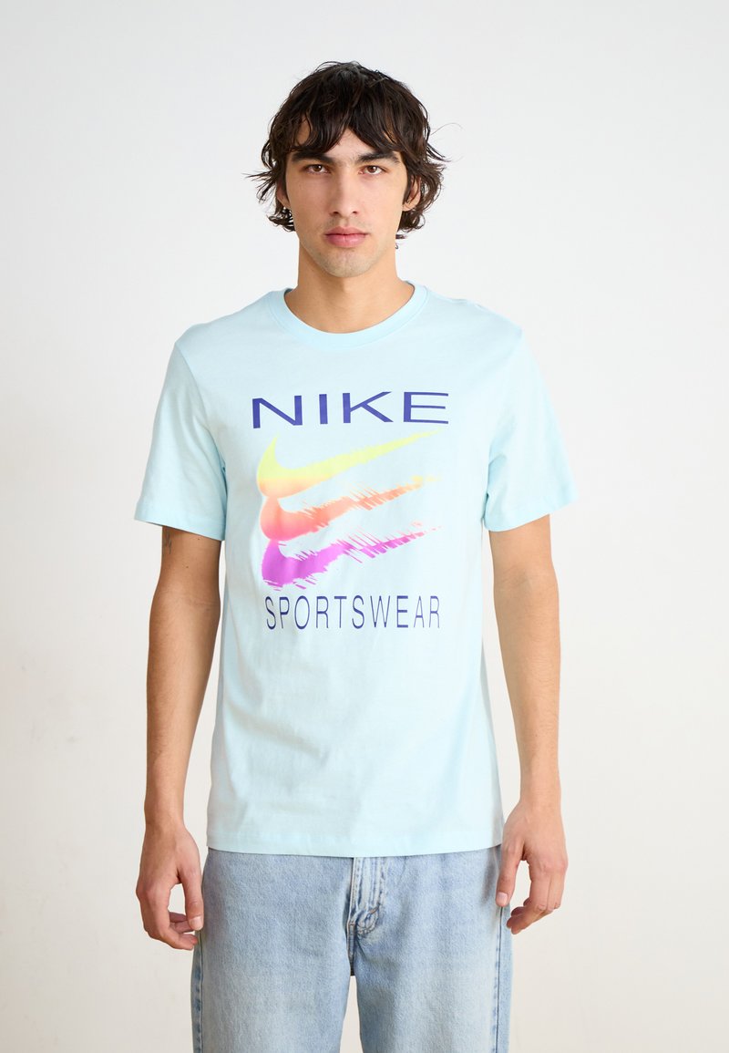 Šviesiai mėlyna medvilninė marškinėliai su dideliu, spalvingu Nike logotipu ir "SPORTSWEAR" tekstu tamsiai violetine spalva, trumpomis rankovėmis ir apvalia iškirpte.