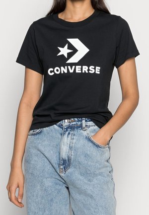 Personne portant un t-shirt noir avec un logo Converse blanc et un jean taille haute bleu clair, une main dans la poche, sur un fond uni.