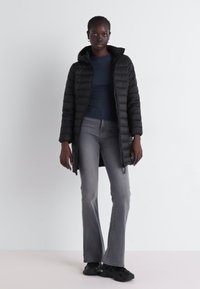 Zwarte gewatteerde jas met kap, doorgestikte ontwerp, ritssluiting, gecombineerd met een donkerblauw aansluitend top en grijze flared jeans, gedragen met zwarte sneakers.