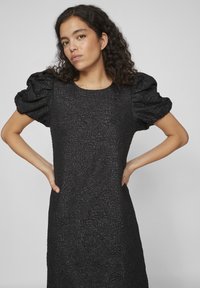 Μαύρο φόρεμα με υφή λουλουδιών, φουσκωτά κοντά μανίκια, χαλαρή εφαρμογή και στρογγυλή λαιμόκοψη. Το ύφασμα φαίνεται παχύ με ανάγλυφες λεπτομέρειες.