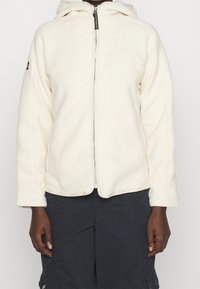 Persona che indossa una giacca in pile bianca con zip frontale e cappuccio, abbinata a pantaloni cargo scuri.