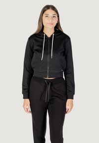 Felpa nera corta con zip frontale, cappuccio e cordini crema. Presenta un tessuto testurizzato e polsini a coste. Abbinata a pantaloni della tuta neri.