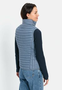 Gepolsterte hellblaue Weste mit hohem Kragen, die horizontale Nähte und eine glatte Textur aufweist, übereinander getragen mit einem dunkelblauen Langarmshirt.