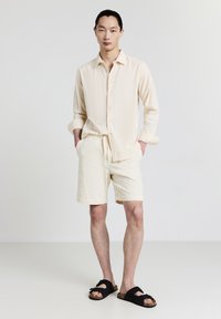 Giovane uomo in piedi con le mani in tasca, indossando una camicia a maniche lunghe crema, pantaloni corti abbinati e sandali neri su uno sfondo bianco semplice.