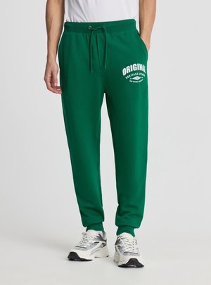 Uomo che indossa pantaloni della tuta verdi con scritta bianca "ORIGINAL Heritage Denim", scarpe da ginnastica bianche e camicia bianca, in piedi con le mani nelle tasche.