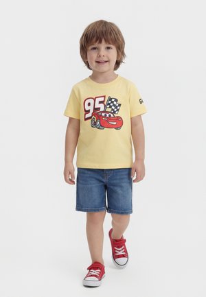 Νεαρός αγόρι περπατάει, φοράει κίτρινη μπλούζα με κόκκινο αγωνιστικό αυτοκίνητο και τον αριθμό 95, τζιν σορτς και κόκκινα αθλητικά παπούτσια με λευκά κορδόνια.