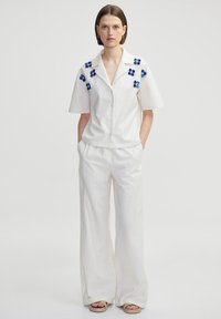 Camisa blanca de manga corta con cierre de botones y bordado floral azul en los hombros, combinada con pantalones anchos y sueltos blancos.