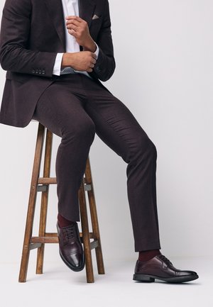 Man seated on wooden stool wearing dark brown suit, white shirt, burgundy socks, and polished dark brown leather shoes against plain background.