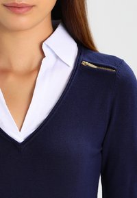 Frau trägt ein weißes Hemd mit Kragen unter einem marineblauen V-Ausschnitt-Pullover mit einem goldenen Reißverschlussdetail auf der Schulter.