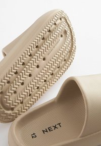 Beige gummisko uden snørebånd med en struktureret overdel og en zigzag-mønstrets sål. Størrelse 12 markeret indeni. "MADE IN CHINA" på bunden.