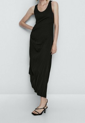 Kobieta ubrana w czarną bezrękawnikową asymetryczną sukienkę midi z plisami i czarne sandały na obcasie z paskami, stojąca na tle jasnej ściany.