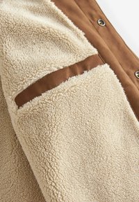 Țesătură de fleece bej cu o suprafață moale, texturată; prezintă un panou maro și nasturi snap vizibili pe margine.
