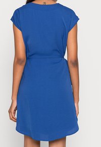 Krátké modré šaty s volným střihem, krátkými rukávy, staženým pasem a mírně zaobleným lemem. Vyrobeny z měkké, texturované látky.
