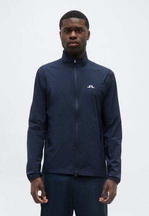 Mlad moški, ki nosi mornarsko modro jakno z zadrgo in visokim ovratnikom, stoji pred preprostim svetlo sivim ozadjem.