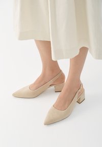 Beige semskede slingback-pumps med spiss tå og blokkheel. Designet har utskjæringer på sidene og en gullspenne som dekorasjon.