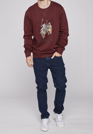 Uomo che indossa una felpa color bordeaux con una grafica di due giocatori di polo a cavallo, jeans blu scuro e scarpe da ginnastica bianche, in piedi con una mano in tasca.