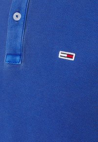 Polo bleu texturé avec un bouton blanc et un petit logo rectangulaire rouge, blanc et noir brodé sur la poitrine.