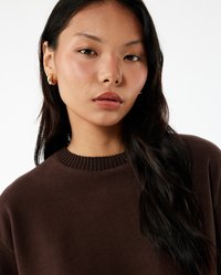 Bruine gestructureerde trui met een geribde hals; het model heeft lang, donker haar en draagt kleine gouden oorringen.