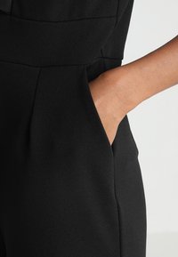 Svart jumpsuit med sidoficka, tillverkad av mjukt tyg med en slät yta och en strukturerad design som framhäver rena linjer och skräddarsydd passform.