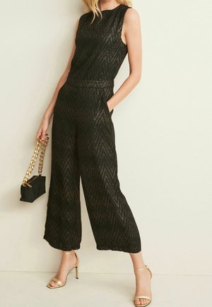 Femme portant une combinaison noire en dentelle sans manches avec un pantalon à jambes larges, tenant une petite pochette noire à chaîne dorée, et des sandales à talons hauts dorées.