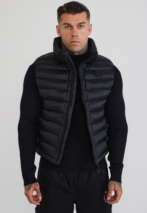 Uomo con capelli corti e scuri che indossa un gilet imbottito nero sopra una maglia nera a maniche lunghe, in piedi davanti a uno sfondo chiaro e uniforme.