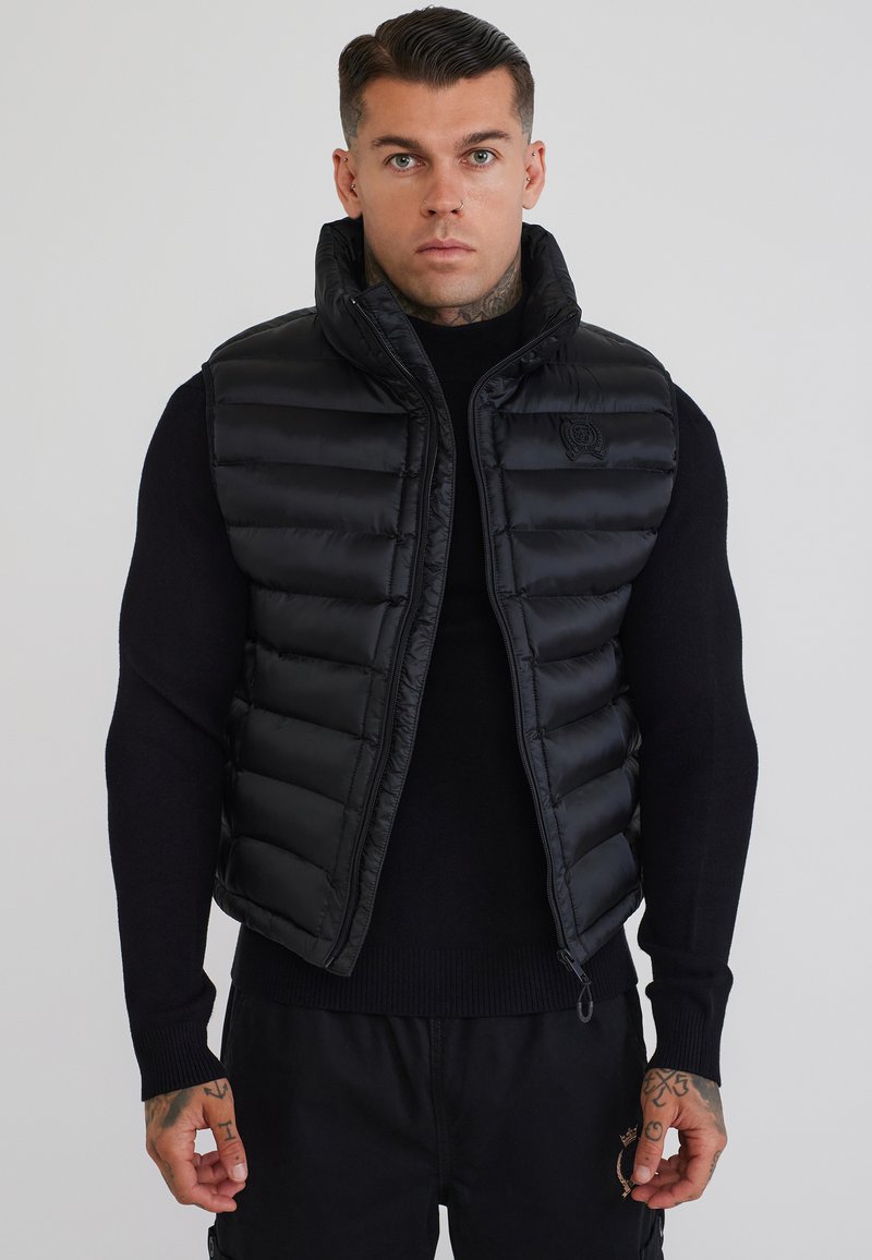 Homme aux cheveux courts et foncés portant un gilet matelassé noir sur une chemise noire à manches longues, debout devant un fond clair uni.