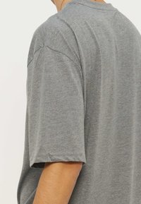 Άτομο με απλό γκρι κοντομάνικο πουκάμισο, εμφανιζόμενο από την πλάτη και τον ώμο, πάνω σε λευκό φόντο.