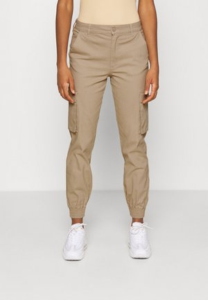 Persona con pantalones cargo beige con puños elásticos en los tobillos y zapatillas blancas, de pie frente a un fondo liso.