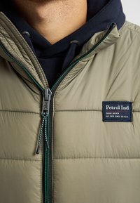Hellgrüne Puffjacke mit strukturiertem Obermaterial, ausgestattet mit einem schwarzen Reißverschluss mit grünen Akzenten und einem Markenzeichen auf der oberen linken Seite.